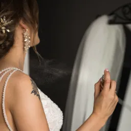 Panna młoda w sukni ślubnej z perłowymi ramiączkami spryskuje się perfumami przed lustrem, widoczny tatuaż róży na ramieniu i ozdobna spinka we włosach.