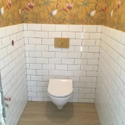 Wnętrze łazienki po remoncie: białe, prostokątne płytki na ścianach do połowy wysokości, nad nimi tapeta w kolorze musztardowym z motywem żurawi i kwiatów, drewnopodobna podłoga, wisząca toaleta...