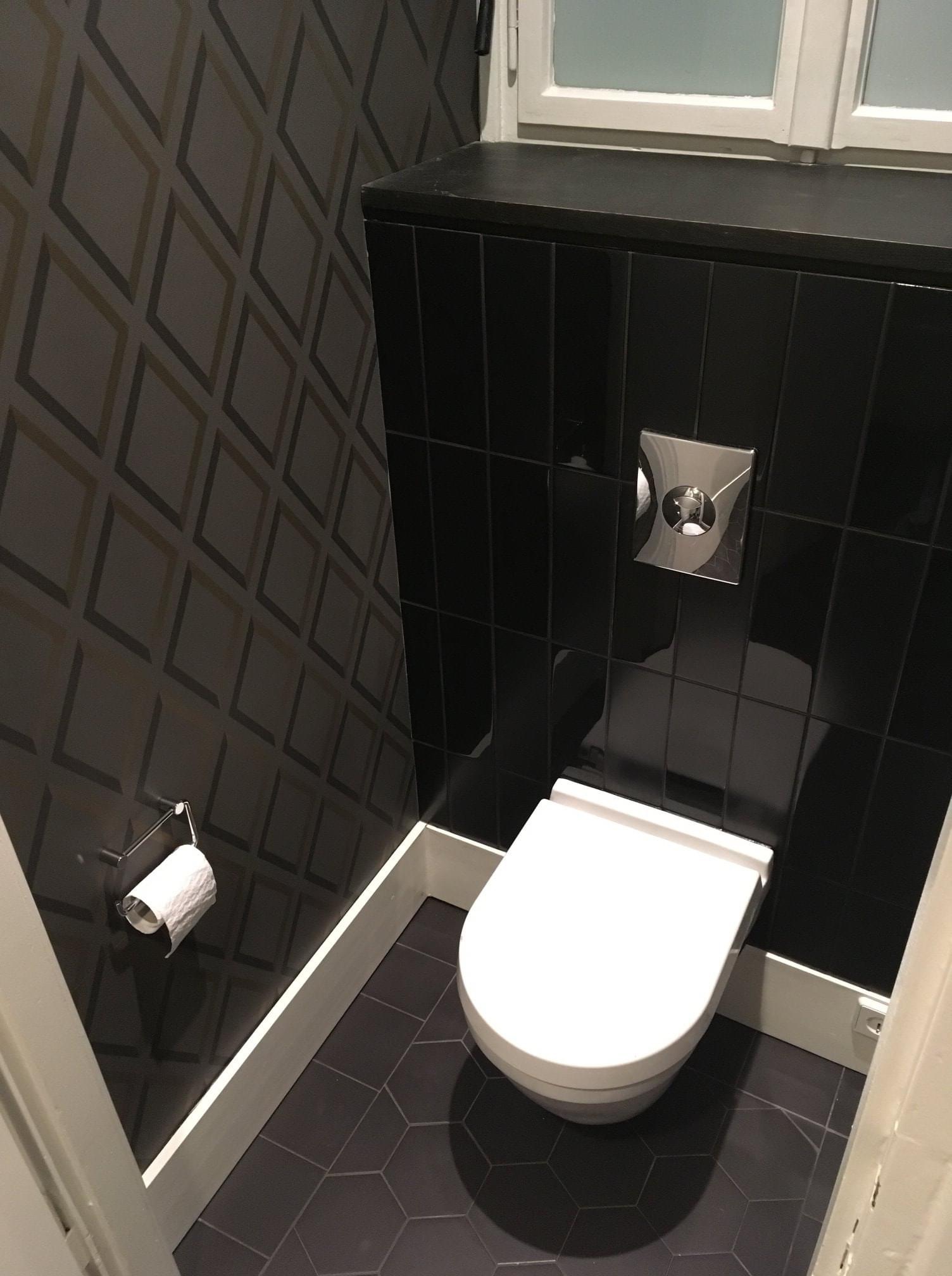 Nowoczesna łazienka w odcieniach szarości i czerni z geometryczną tapetą, czarnymi płytkami i białą toaletą.