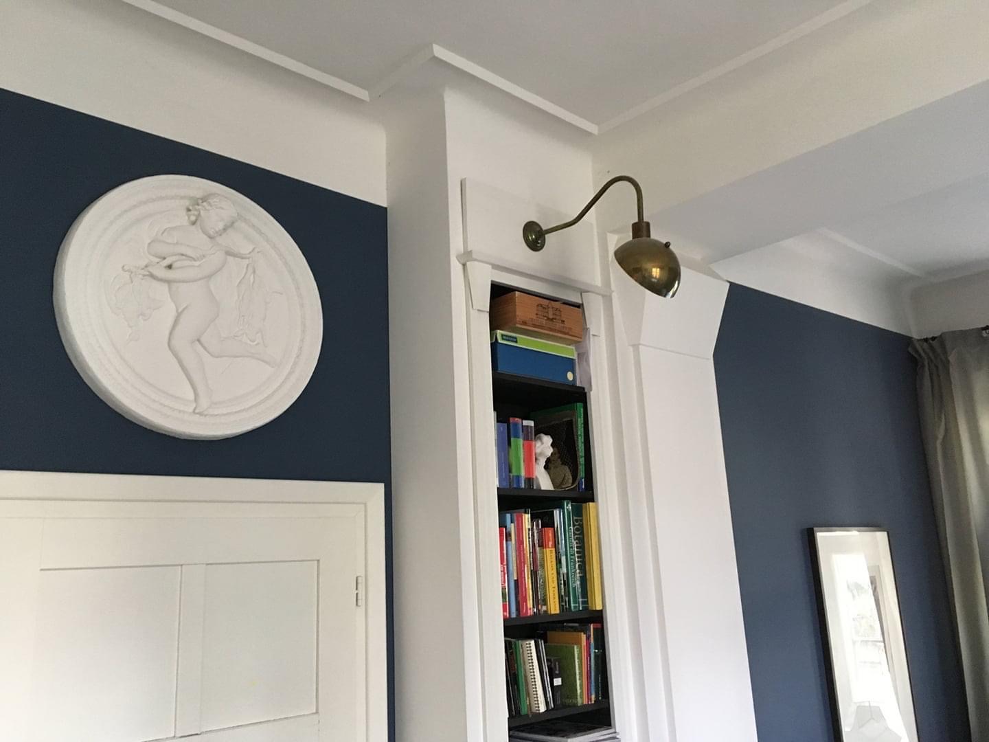 Granatowa ściana z białym, okrągłym płaskorzeźbą przedstawiającą postać, wbudowany regał z książkami oświetlony mosiężną lampą, białe listwy przysufitowe i drzwi.