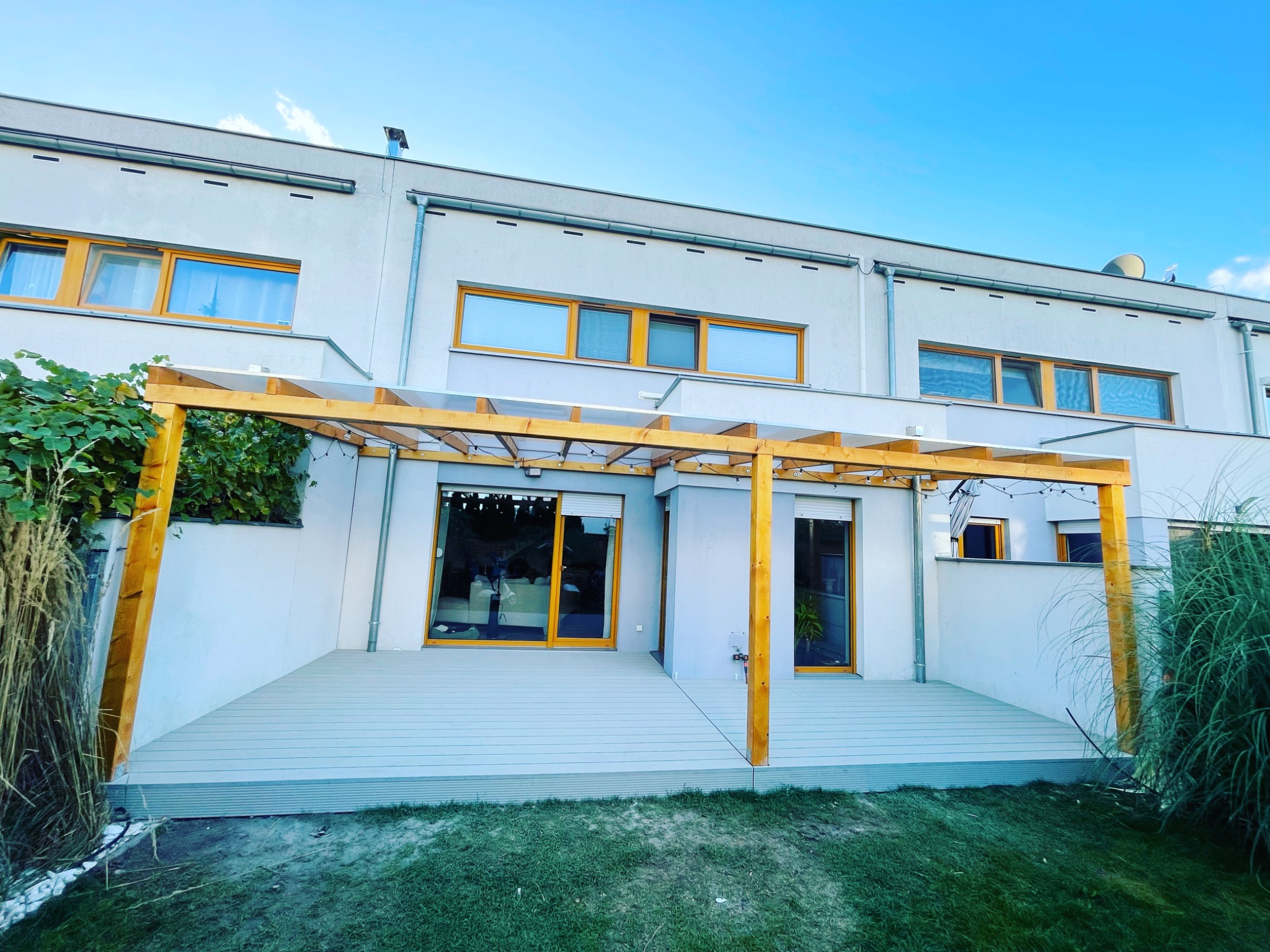 Nowoczesny dom szeregowy z szarą kompozytową podłogą i drewnianą pergolą, oświetloną girlandą żarówek, widok z niskiej perspektywy na ogród.