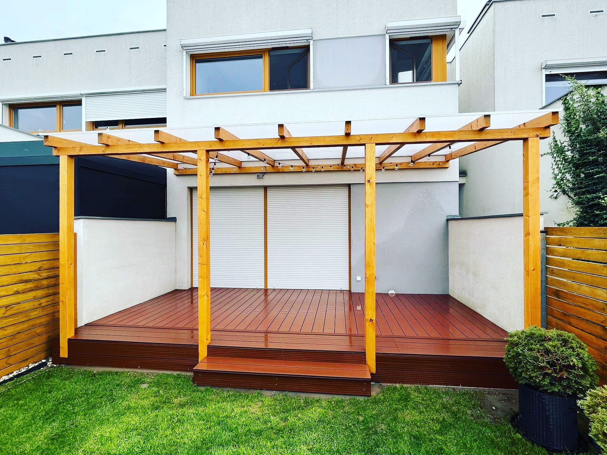 Nowoczesny taras kompozytowy z drewnianą pergolą i trzema schodkami, otoczony zadbanym trawnikiem i ogrodzeniem z desek.