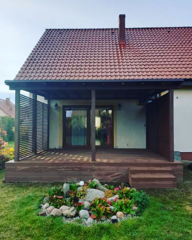 Zadaszony taras z brązowych desek z niewielkimi schodkami i dekoracyjnym skalniakiem u podstawy, przylegający do domu z czerwoną dachówką.