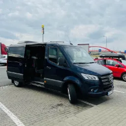 Ciemny bus Mercedes Sprinter z otwartymi drzwiami przesuwnymi na parkingu z kostki brukowej; w tle widoczny czerwony samochód osobowy z bagażnikiem dachowym, McDonald's i wiadukt.
