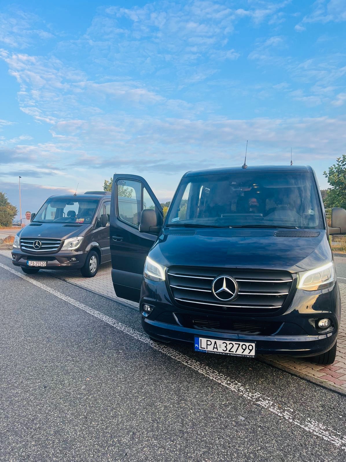 Dwa busy Mercedes-Benz Sprinter na parkingu, jeden czarny z otwartymi drzwiami, drugi szary, błękitne niebo w tle.