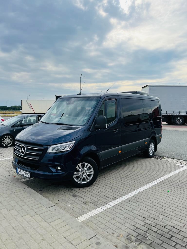 Ciemnoniebieski bus Mercedes-Benz Sprinter, zaparkowany na parkingu z kostki brukowej, widoczny z boku, z lekkiego ukosa, na tle pochmurnego nieba i zaparkowanych ciężarówek.