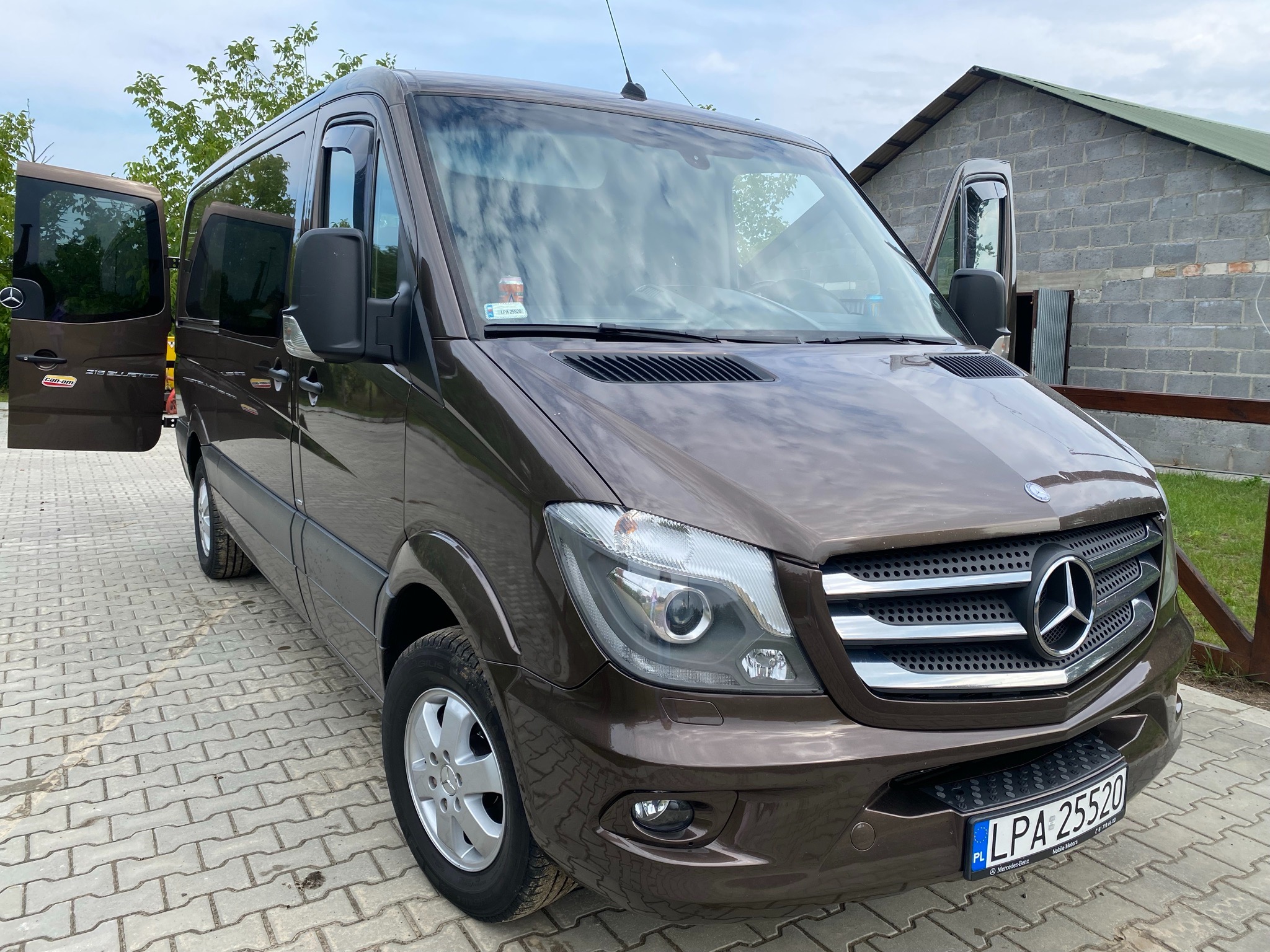 Brązowy bus Mercedes-Benz Sprinter z otwartymi drzwiami bocznymi, zaparkowany na kostce brukowej przed budynkiem z szarej cegły.