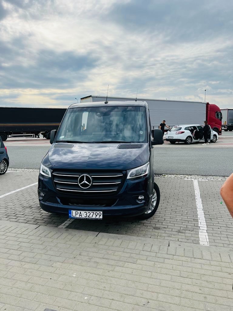 Ciemny bus Mercedes z polską rejestracją LPA 32799 zaparkowany na parkingu z kostki brukowej, w tle inne pojazdy i osoby przy białym samochodzie osobowym.