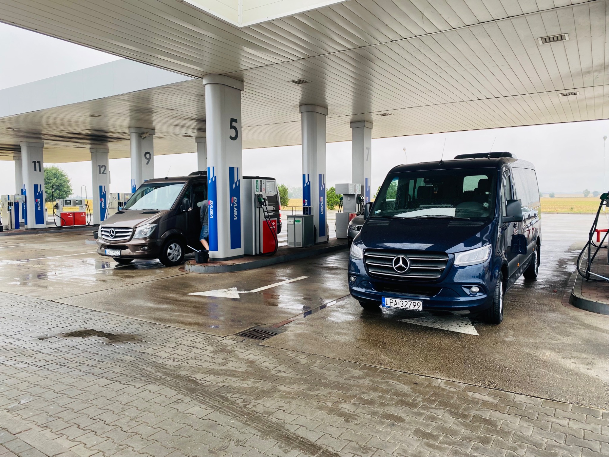 Dwa busy marki Mercedes-Benz Sprinter, jeden granatowy z przodu, drugi brązowy w tle, stoją na stacji benzynowej pod zadaszeniem, z widocznymi dystrybutorami paliwa i numerami stanowisk.