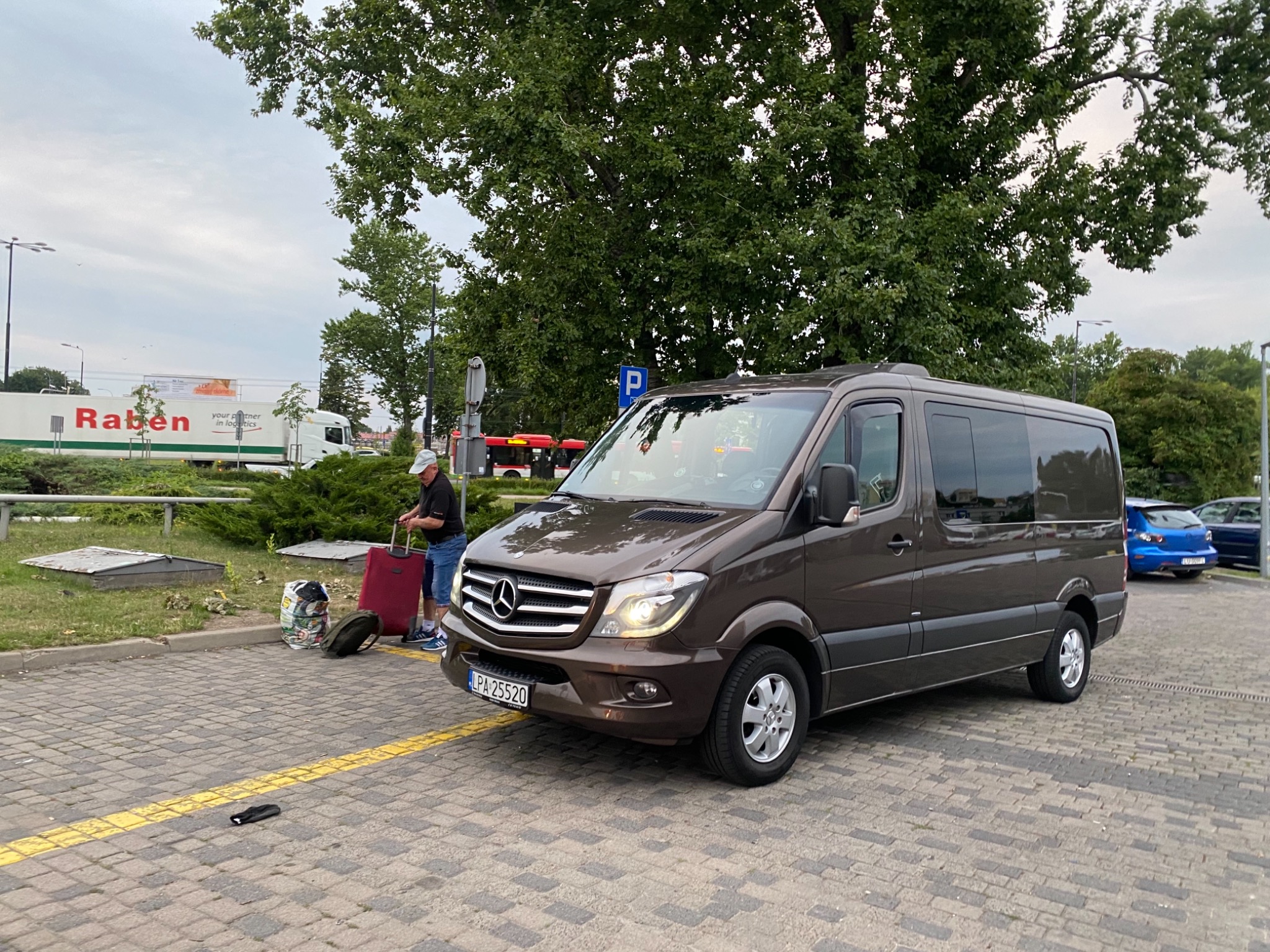 Brązowy bus Mercedes-Benz Sprinter zaparkowany na brukowanym placu, obok stoi mężczyzna z czerwoną walizką i torbami, w tle widoczny budynek i zieleń.