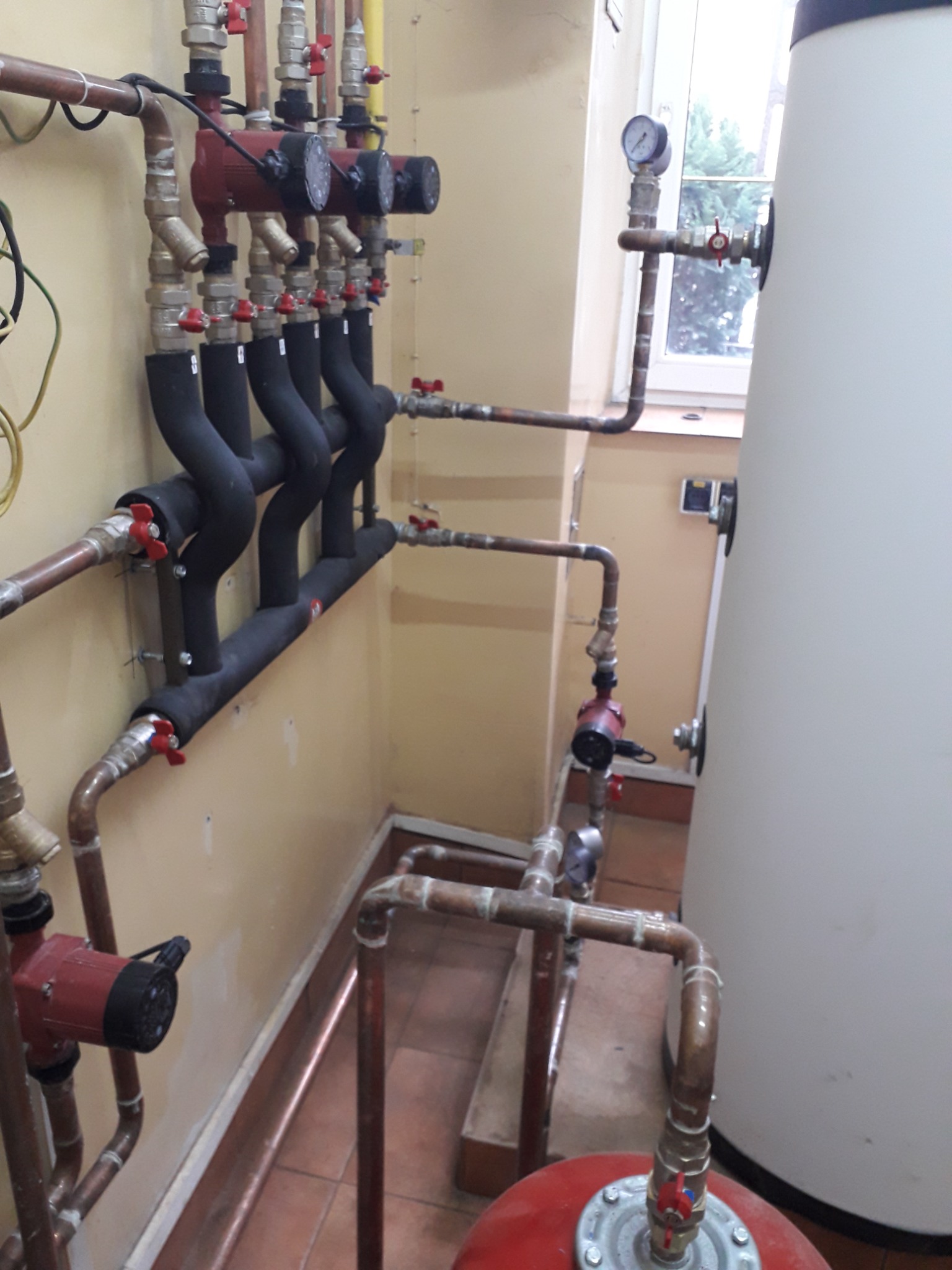 Instalacja grzewcza w kotłowni: widoczne pompy obiegowe z izolacją termiczną, manometry, zawory kulowe z czerwonymi uchwytami, rury miedziane oraz biały zasobnik ciepłej wody użytkowej przy oknie.