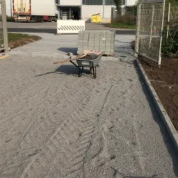 Przygotowanie podłoża z tłucznia pod układanie kostki brukowej, widoczna taczka z narzędziami i paleta z kostką, w tle hala magazynowa