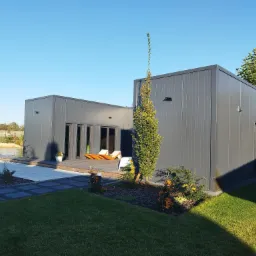 Nowoczesny budynek modułowy z szarych płyt warstwowych, otoczony zielonym trawnikiem i ozdobnymi krzewami, z tarasem i leżakami na zewnątrz.