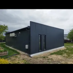 Gotowa hala z ciemnych płyt warstwowych, z dwoma oknami, z klimatyzatorami na ścianie bocznej, widok z zewnątrz na tle pochmurnego nieba i ogrodu.