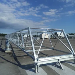 Metalowa konstrukcja nośna, prawdopodobnie pod panele słoneczne lub klimatyzację, w trakcie montażu na płaskim dachu budynku przemysłowego, widok z g&oacute;ry, błękitne niebo z chmurami w tle.