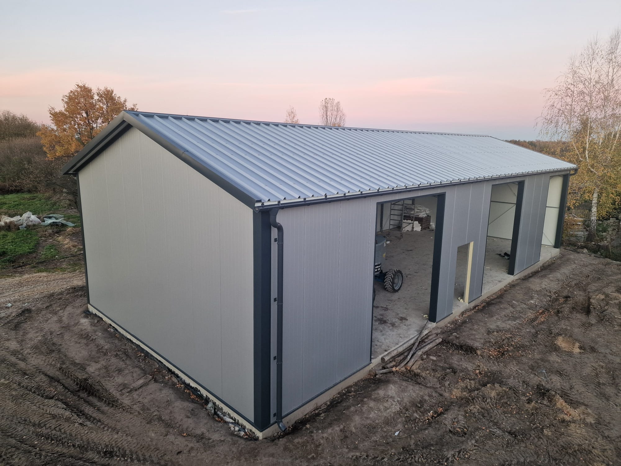 Nowoczesna hala stalowa z szarej płyty warstwowej i stalową konstrukcją, widok z zewnątrz, częściowo otwarte wrota, teren wokół niezagospodarowany, jesienne drzewa w tle, wieczorne niebo.