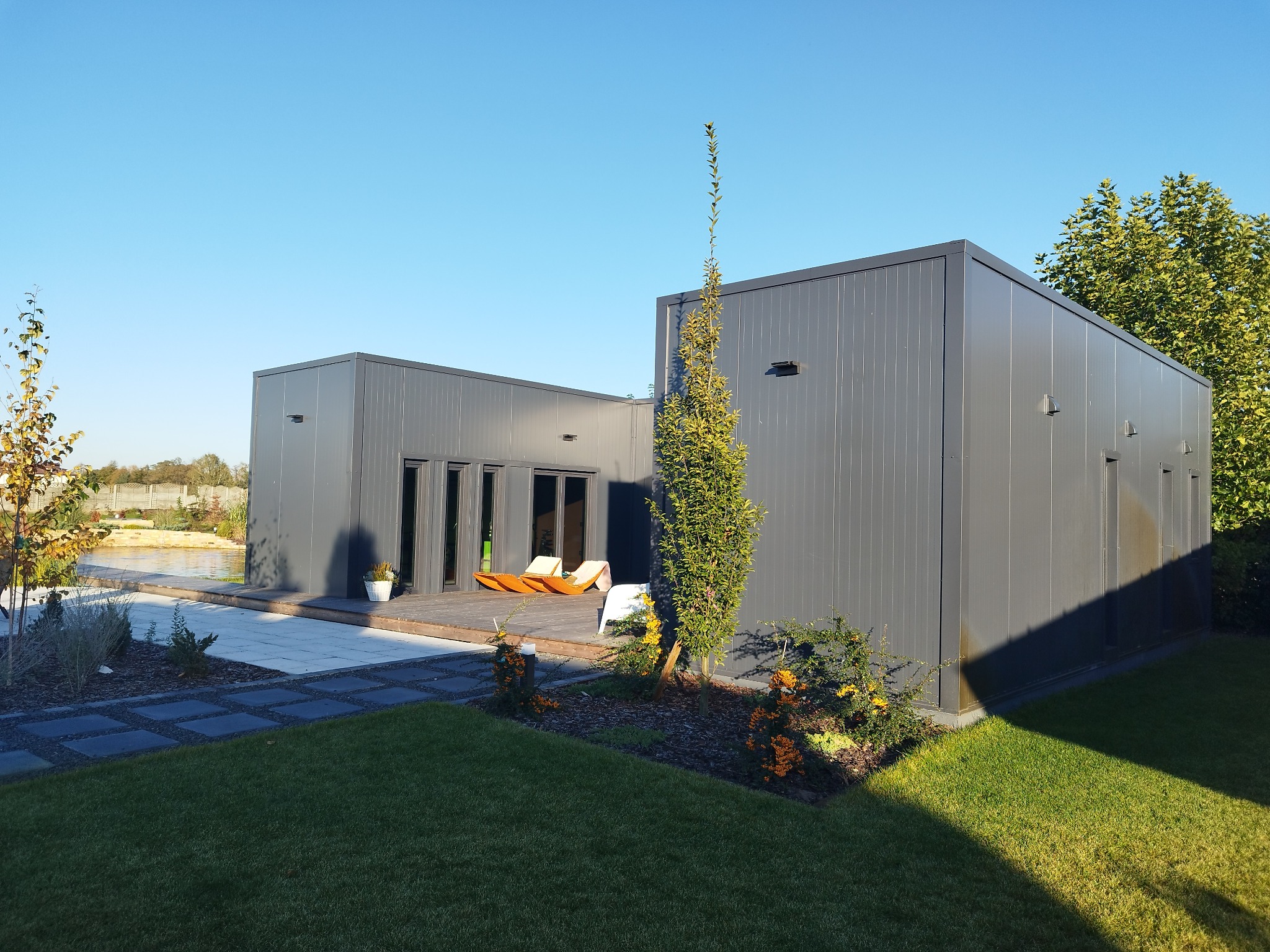 Nowoczesny budynek modułowy z szarych płyt warstwowych, otoczony zielonym trawnikiem i ozdobnymi krzewami, z tarasem i leżakami na zewnątrz.
