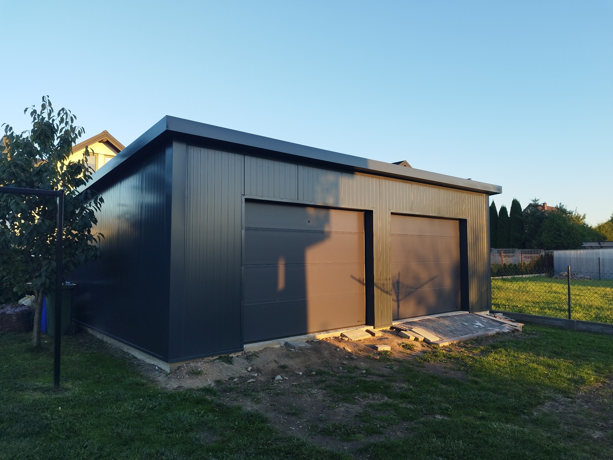 Gotowa, dwustanowiskowa hala garażowa z szarej blachy trapezowej, z dwoma bramami segmentowymi, usytuowana na trawniku.