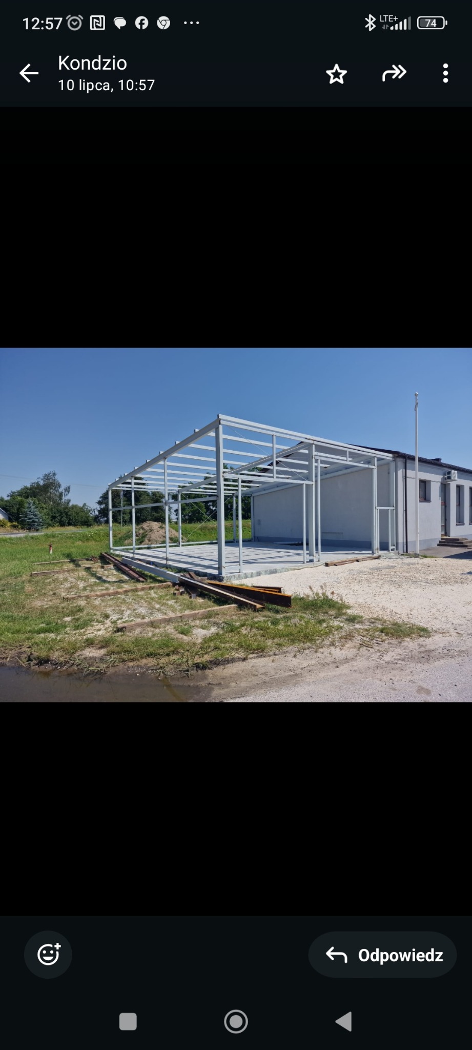 Stalowa konstrukcja hali w trakcie budowy, widoczne elementy szkieletu na betonowej podstawie, w tle budynek z białymi ścianami i błękitne niebo.