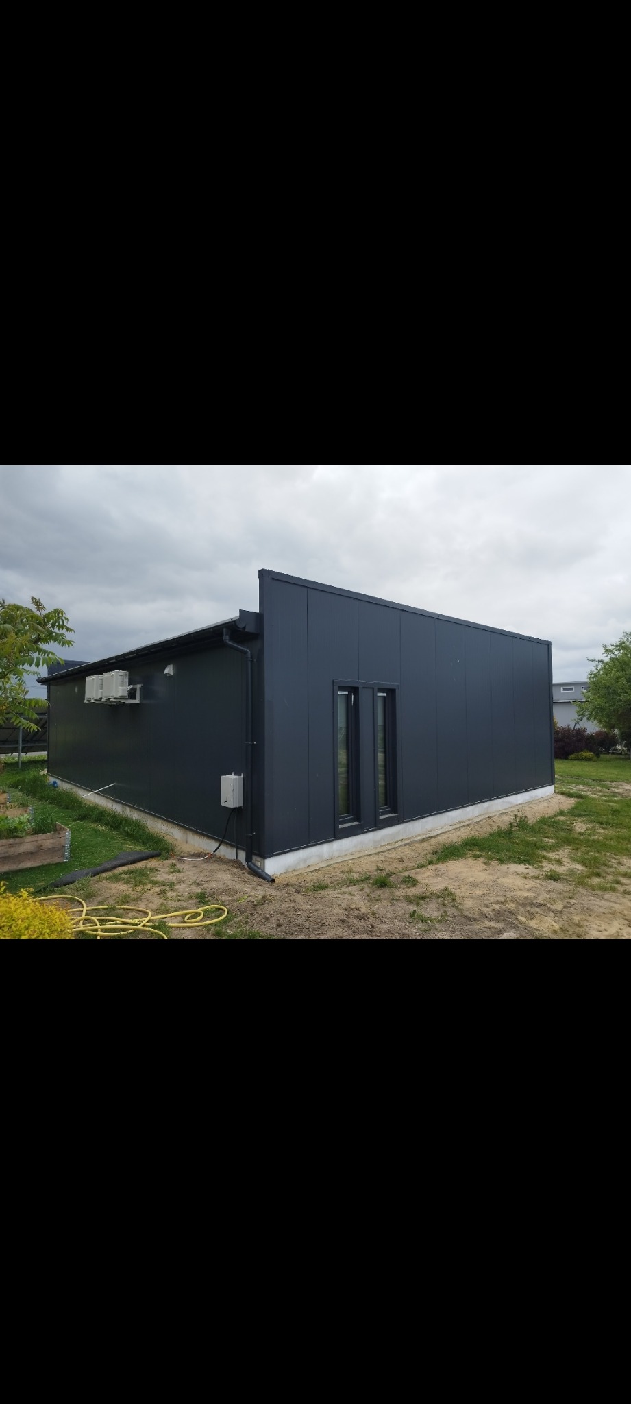 Gotowa hala z ciemnych płyt warstwowych, z dwoma oknami, z klimatyzatorami na ścianie bocznej, widok z zewnątrz na tle pochmurnego nieba i ogrodu.