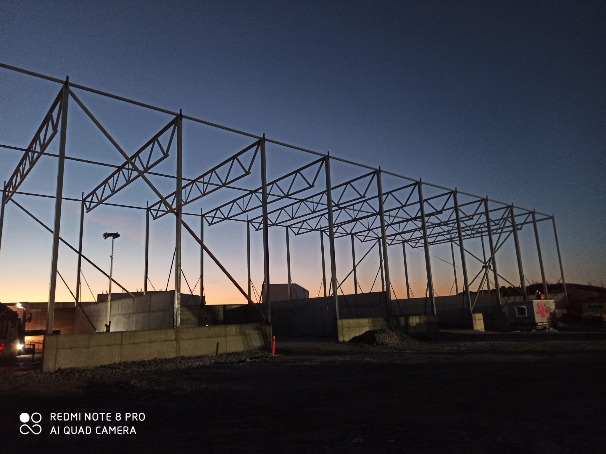Konstrukcja stalowa hali w trakcie budowy, widoczna na tle zachodzącego słońca. Elementy konstrukcyjne zespawane, tworzące szkielet budynku. W tle betonowe ściany i zaparkowany sprzęt budowlany.
