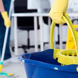 Osoba w żółtych rękawicach trzyma niebieskie wiadro z wyciskaczem do mopa, w tle osoba myjąca podłogę mopem i żółty znak ostrzegawczy.