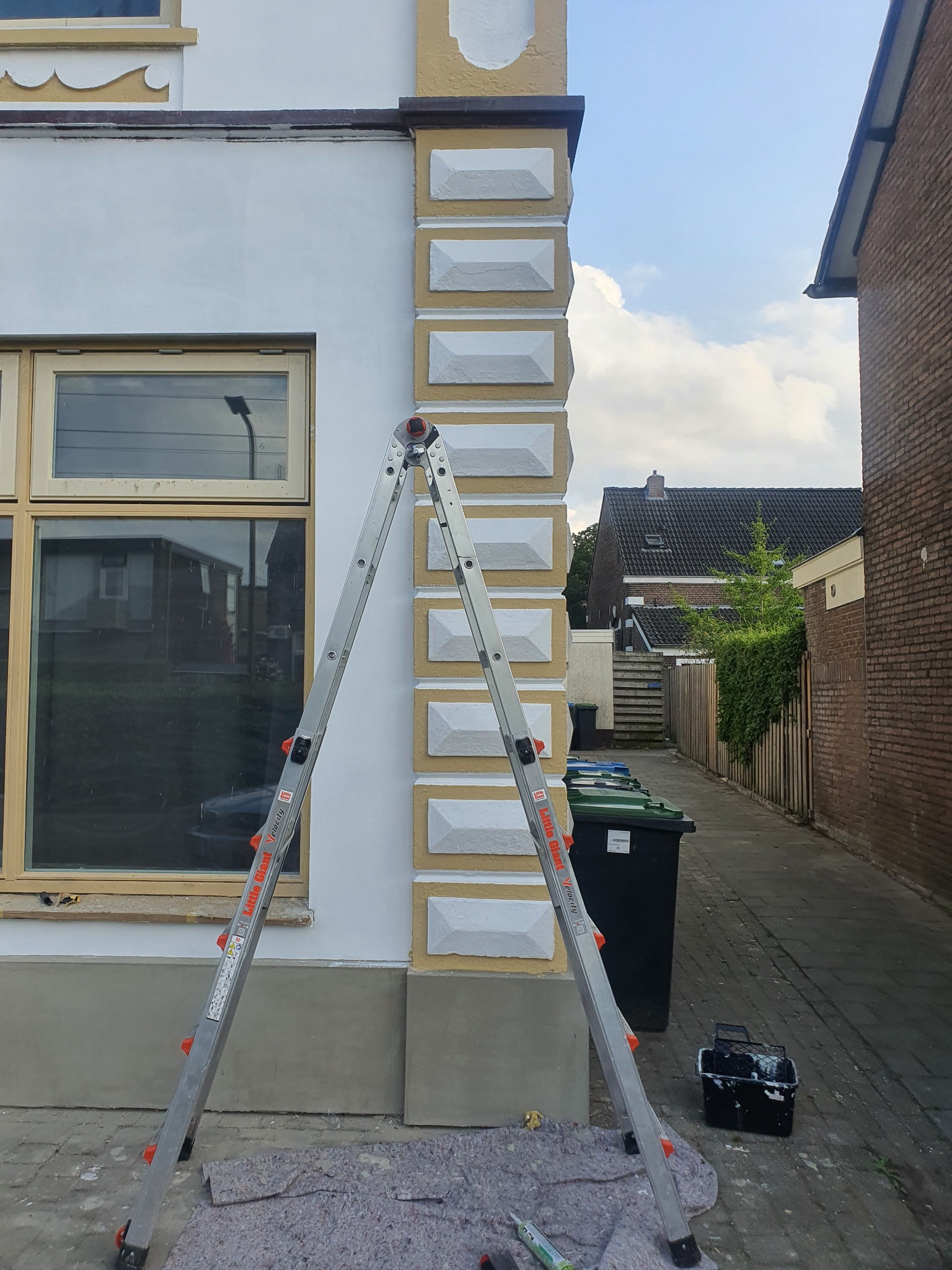 Ściana budynku z dekoracyjnymi elementami pomalowana na biało i żółto, z rozstawioną drabiną Little Giant Velocity, widoczne narzędzia malarskie i pojemnik z farbą, w tle chodnik i budynki sąsiednie.