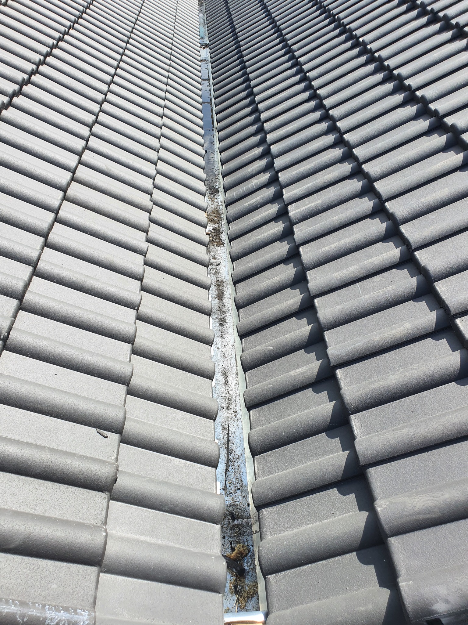 Szare dachówki ceramiczne ułożone rzędami, widoczny fragment rynny z zanieczyszczeniami, perspektywa z góry.
