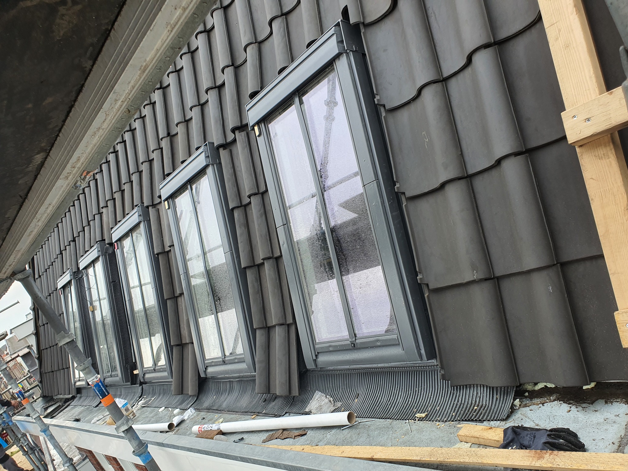 Montaż okien dachowych na krytym czarną dachówką dachu z widocznymi elementami rusztowania i narzędziami pracy.