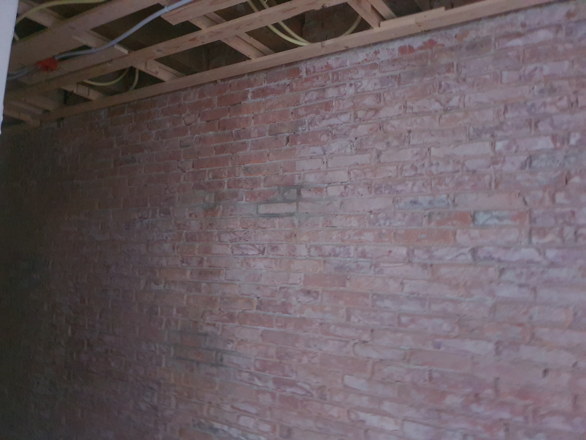 Ceglana ściana wewnętrzna z widoczną konstrukcją drewnianą sufitu i instalacją elektryczną, ukazująca stan przed pracami wykończeniowymi.