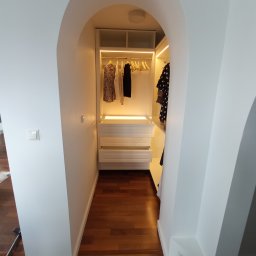 Dakon Daniel Kondzior - Biała szafa wnękowa z oświetleniem LED, widoczna z perspektywy wejścia o łukowatym kształcie, na podłodze parkiet z ciemnego drewna.