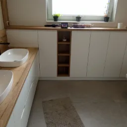 Nowoczesna łazienka z dwoma umywalkami nablatowymi na drewnianym blacie, białymi szafkami podblatowymi z otwartą półką, oświetlonym oknem i beżową podłogą.