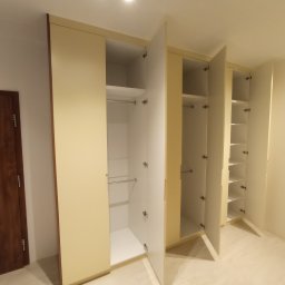 Dakon Daniel Kondzior - Beżowa szafa wnękowa z otwartymi białymi drzwiami, widoczne półki i drążki na ubrania, zamontowana w jasnym pomieszczeniu z drewnianą podłogą i drzwiami.