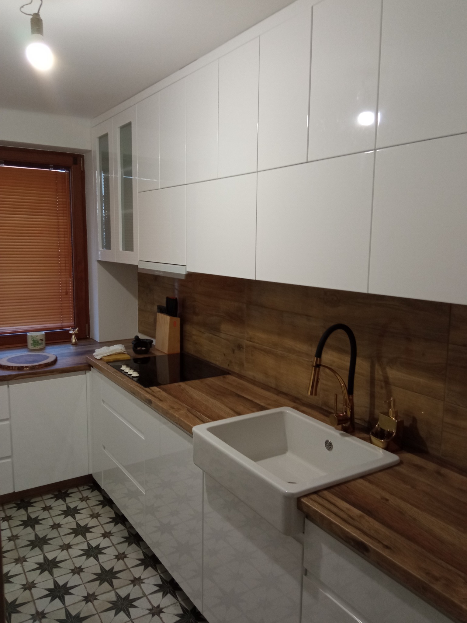 Nowoczesna kuchnia z białymi, błyszczącymi szafkami, drewnianym blatem i ceramicznym zlewem gospodarczym. Uwagę zwraca złota bateria i wzorzysta podłoga.