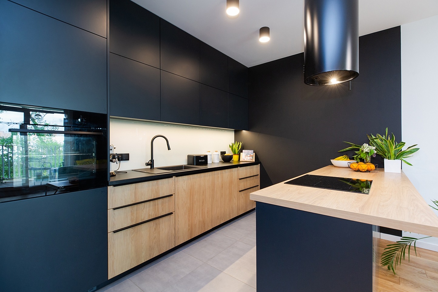 Nowoczesna kuchnia z drewnianymi frontami szafek dolnych i ciemnymi górnymi, czarną armaturą i wyspą z płytą indukcyjną, oświetlona punktowymi lampami sufitowymi.