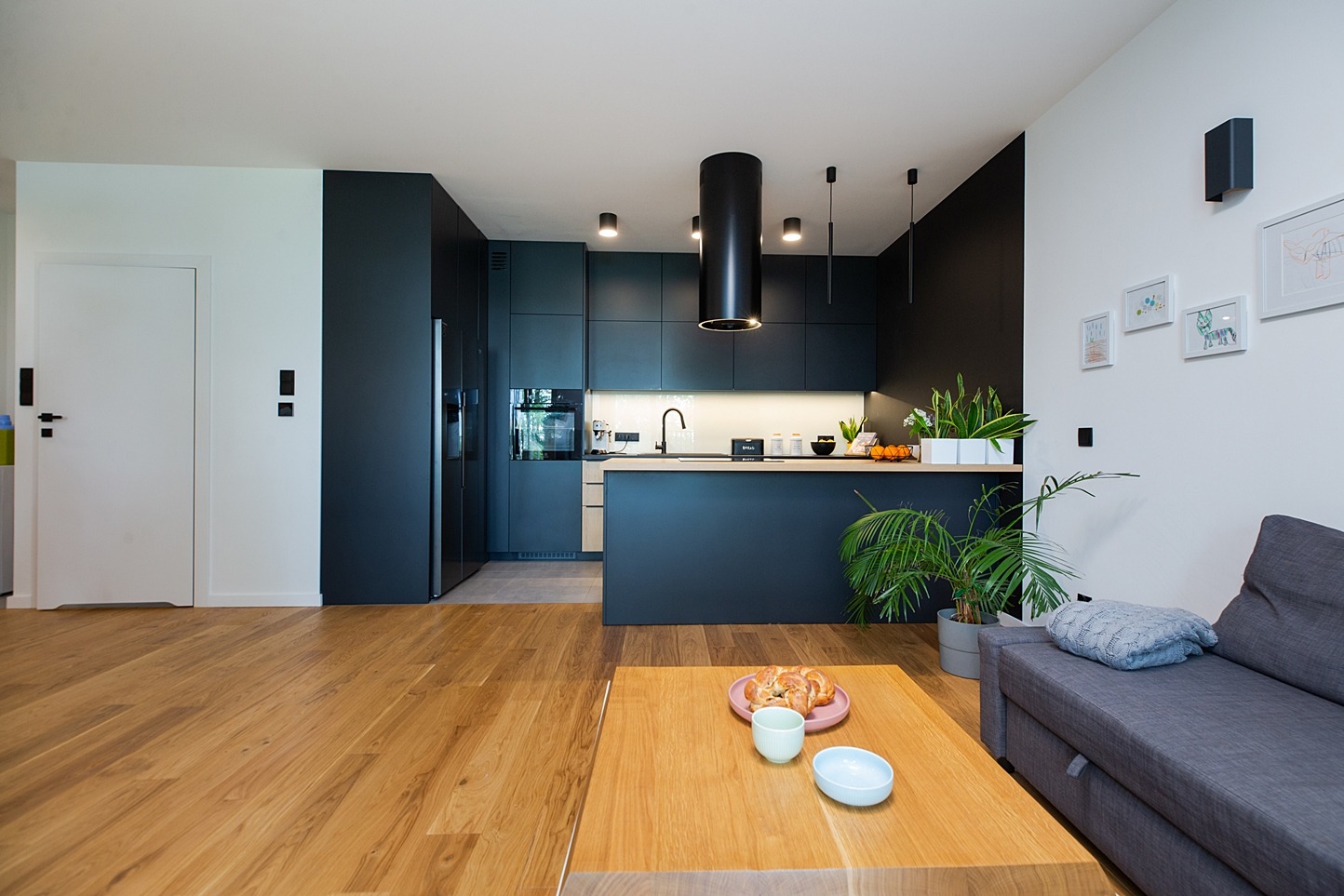 Nowoczesna kuchnia z ciemnymi szafkami i drewnianym blatem, widok z salonu z drewnianą podłogą i stolikiem kawowym z ciastkami.