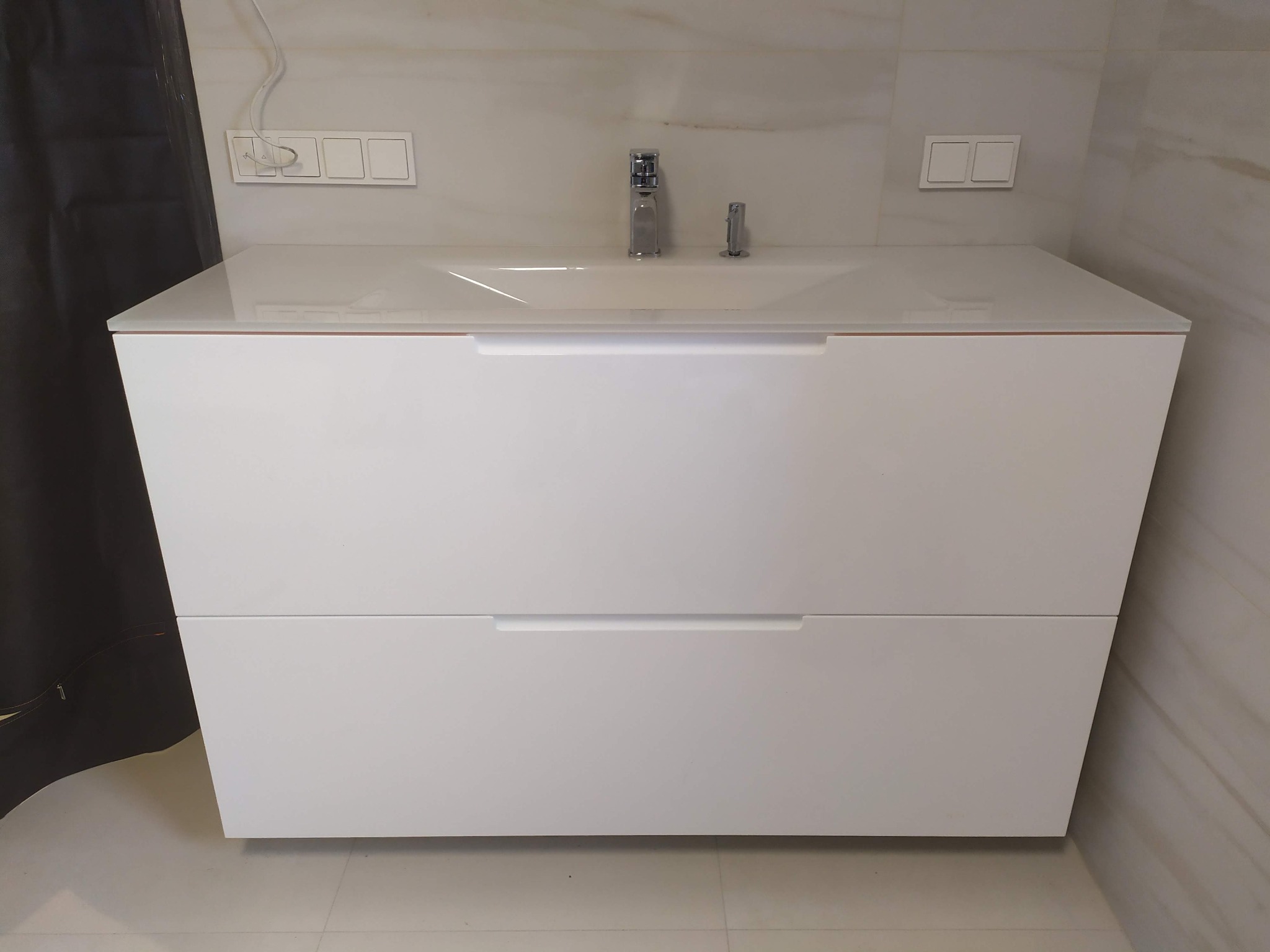Biała szafka łazienkowa z dwiema szufladami i umywalką nablatową, nowoczesny minimalizm, widoczne włączniki światła na ścianie pokrytej marmurkową płytką.