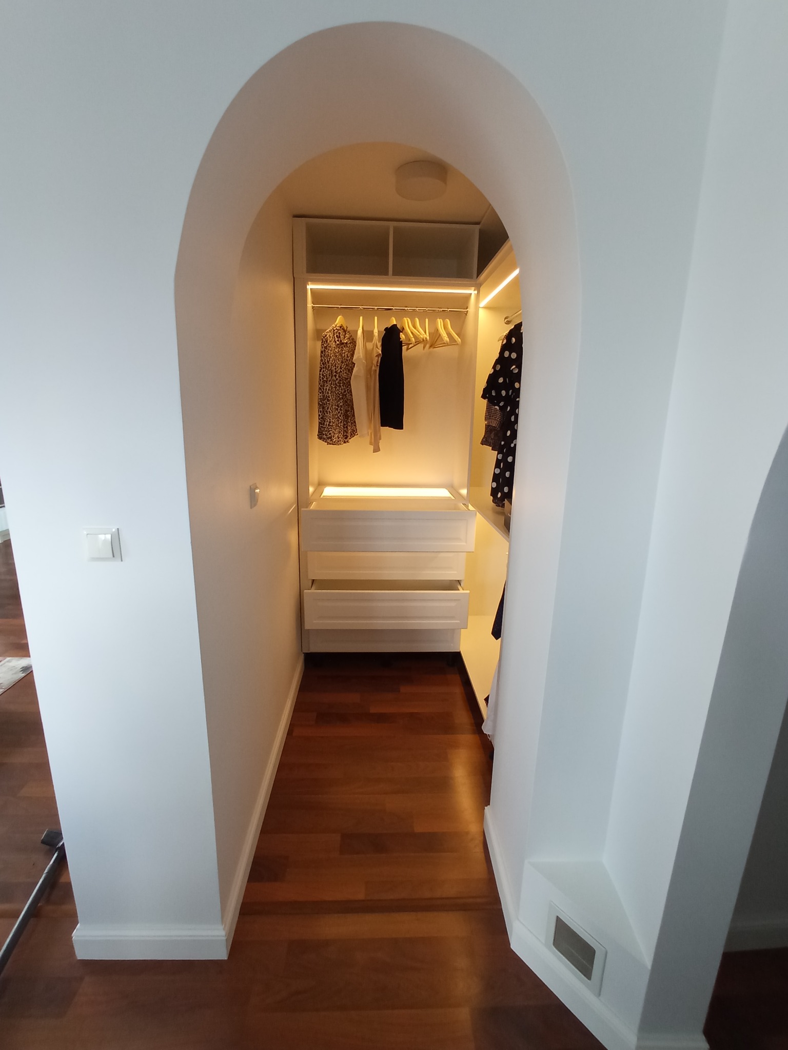 Biała szafa wnękowa z oświetleniem LED, widoczna z perspektywy wejścia o łukowatym kształcie, na podłodze parkiet z ciemnego drewna.