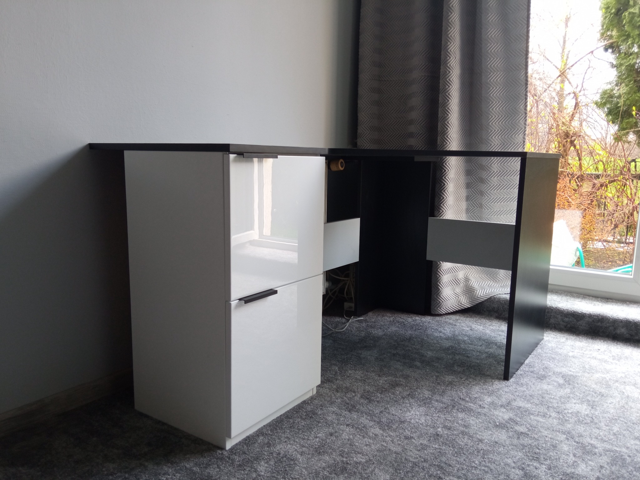 Nowoczesne biurko z czarnym blatem i białymi szafkami z połyskiem, widoczne okno z zasłonami w tle.