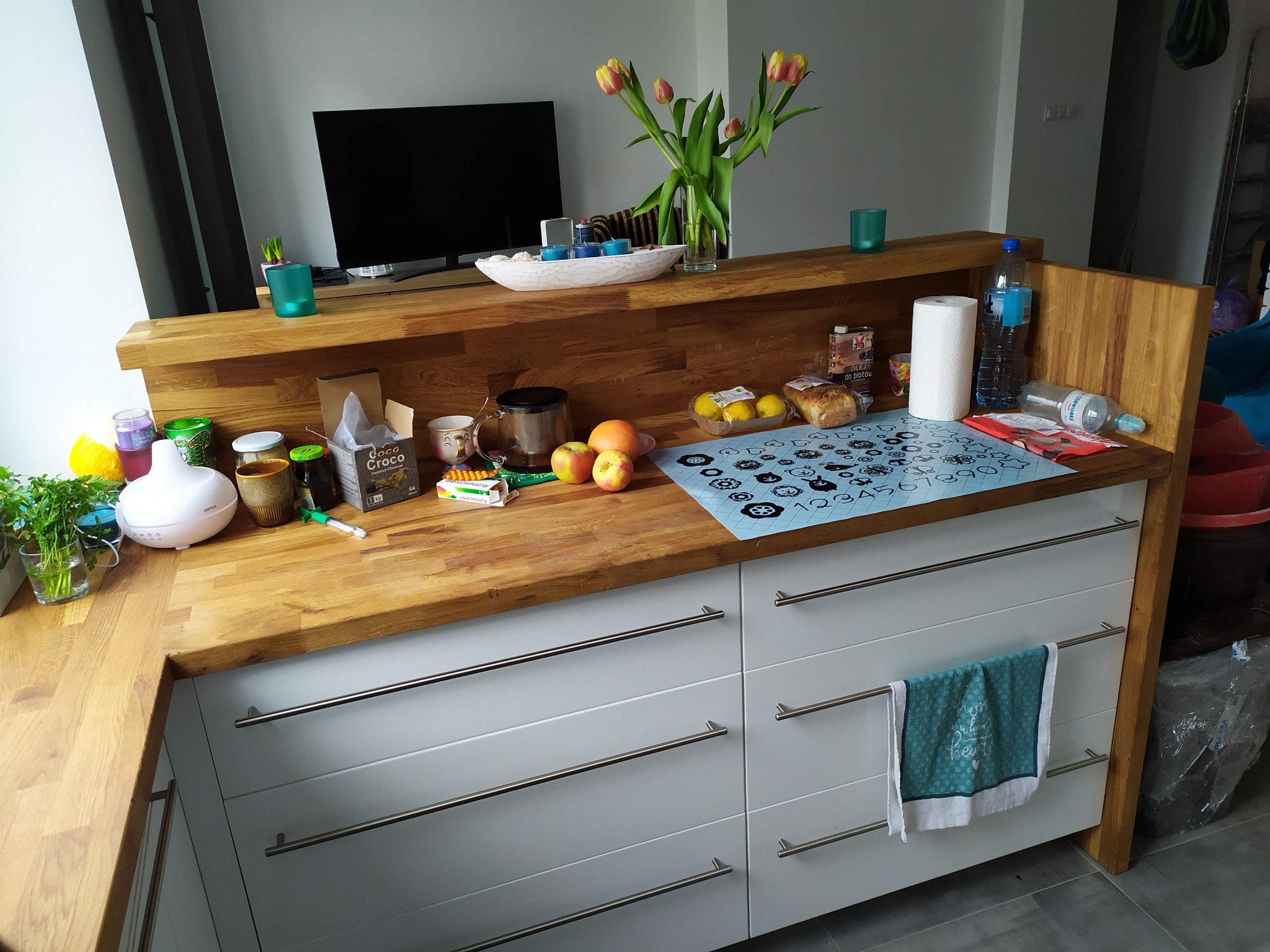 Drewniany blat kuchenny z białymi frontami szuflad, na którym znajdują się owoce, przyprawy i akcesoria kuchenne, w tle telewizor i bukiet tulipanów.