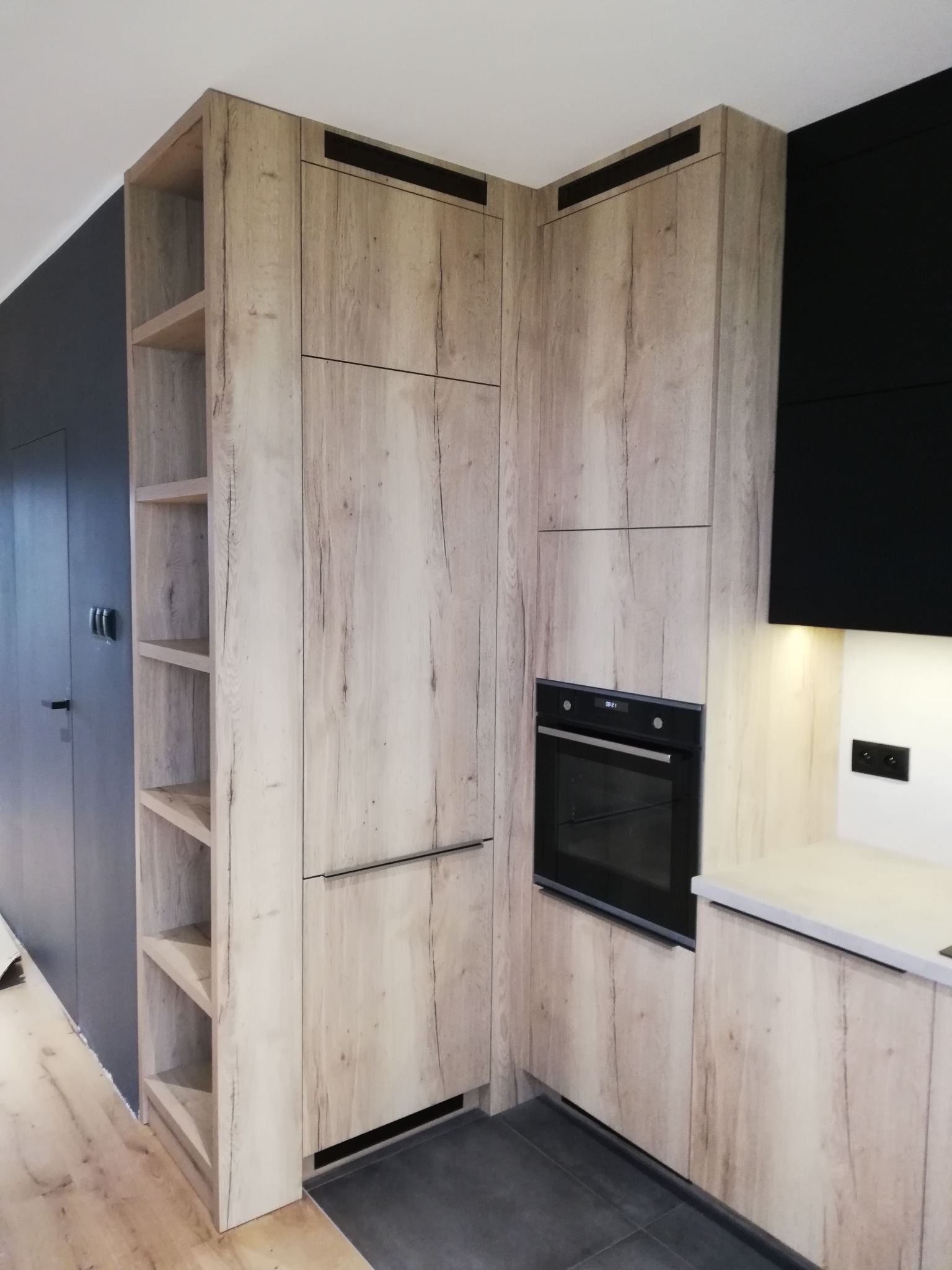 Jasna kuchnia z drewnianymi frontami, czarny piekarnik w zabudowie, otwarte półki z boku, ciemnoszara ściana w tle.