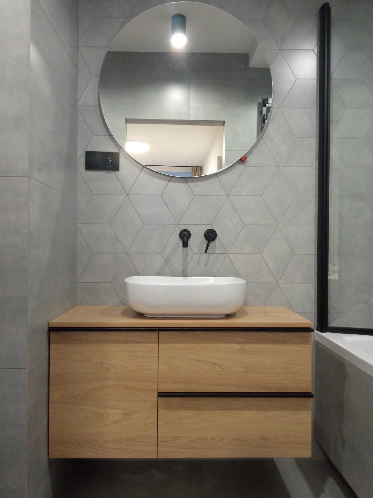 Nowoczesna łazienka z drewnianą szafką pod umywalką, okrągłym lustrem i szarą geometryczną mozaiką na ścianie.