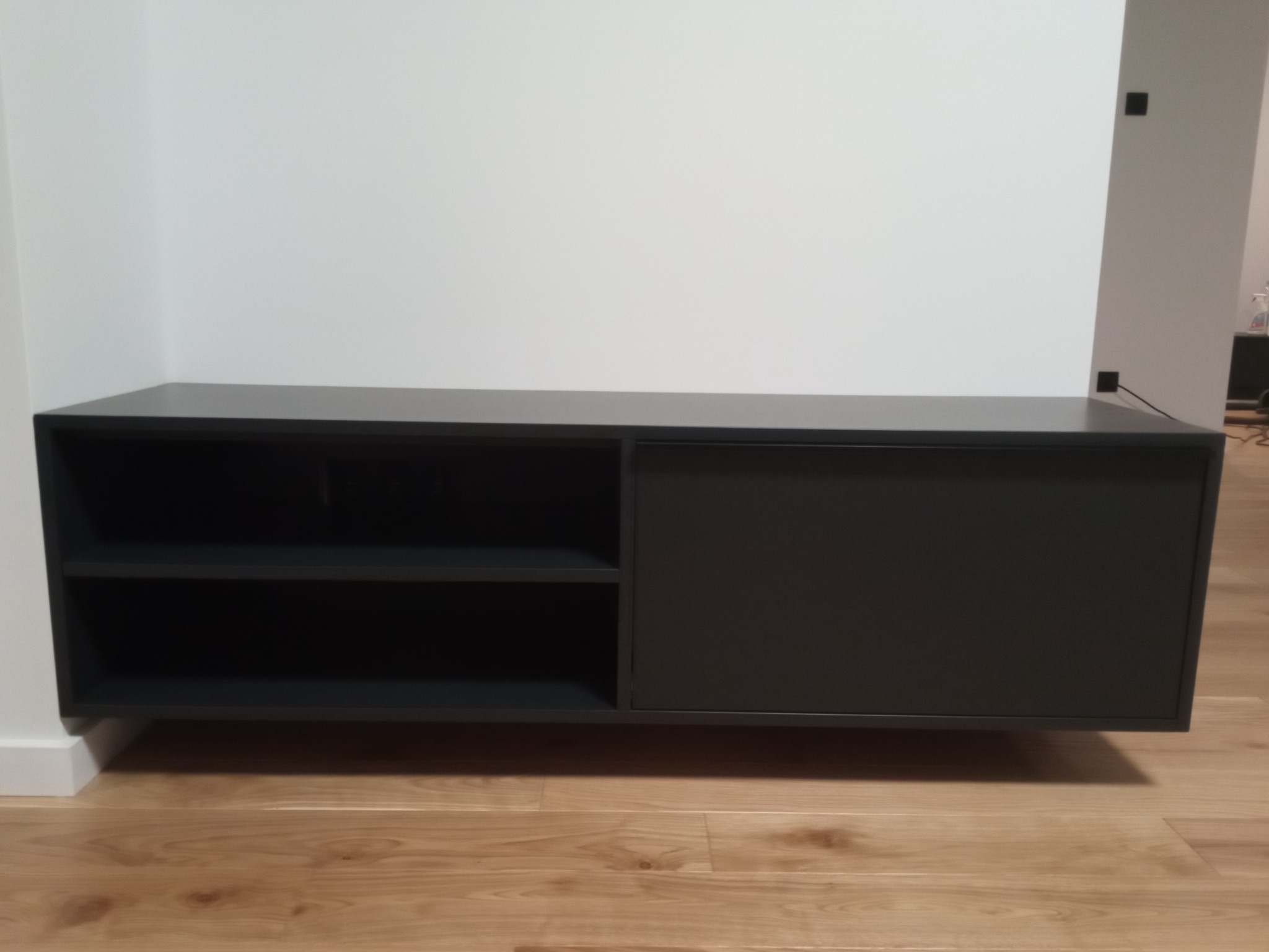 Ciemnoszara szafka RTV zawieszona na ścianie, z przesuwanymi drzwiami i otwartymi półkami, na tle białej ściany i drewnianej podłogi.