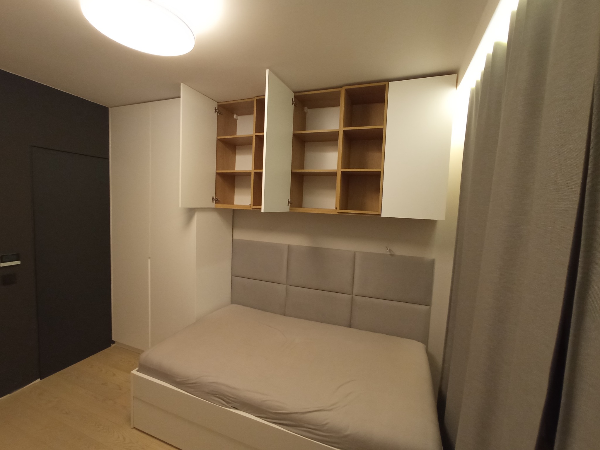 Nowoczesna sypialnia z zabudową meblową: białe szafy z otwartymi półkami nad łóżkiem z szarym zagłówkiem, minimalistyczny design.