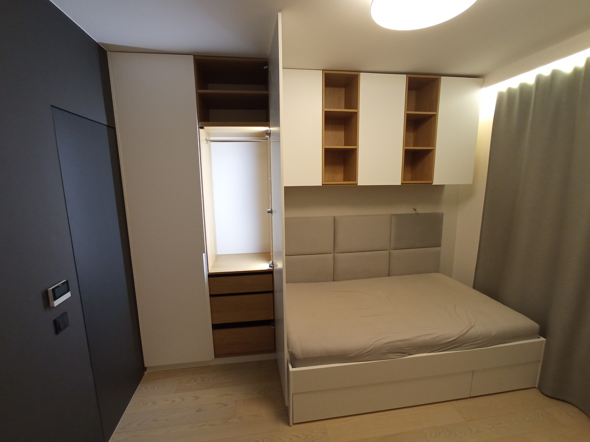 Nowoczesna sypialnia z zabudową meblową: szafa wnękowa z drewnianymi szufladami, białe szafki wiszące z otwartymi półkami z jasnego drewna oraz łóżko z tapicerowanym zagłówkiem w odcieniach beżu.