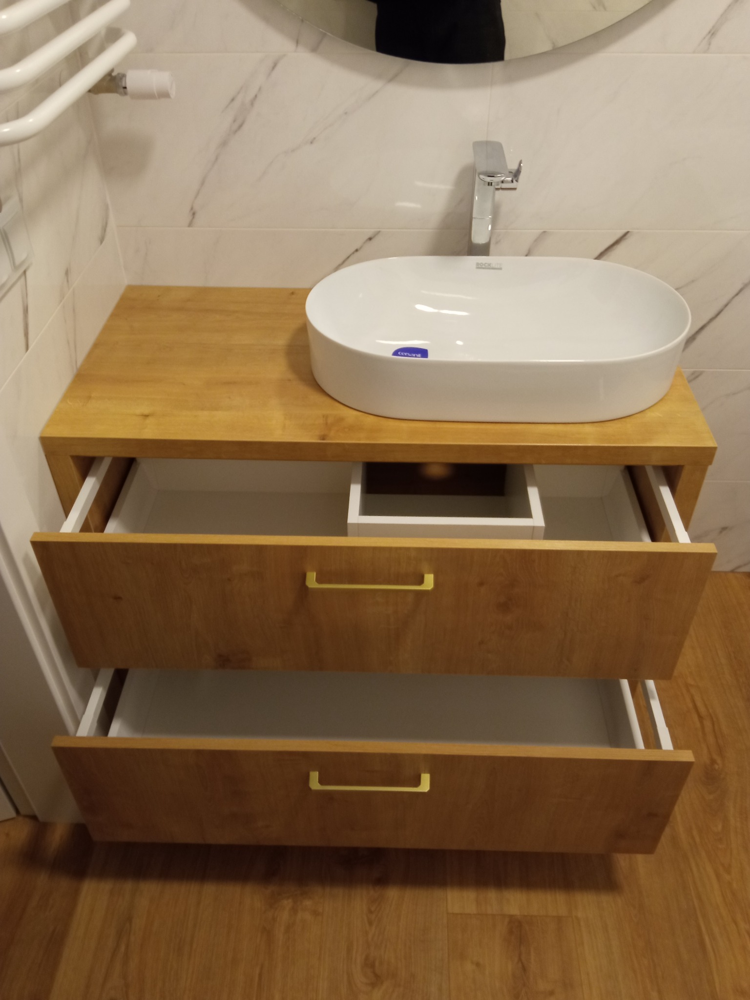 Biała umywalka nablatowa na szafce z dwiema szufladami z jasnego drewna, złote uchwyty, widoczne wnętrze szuflad i armatura łazienkowa.