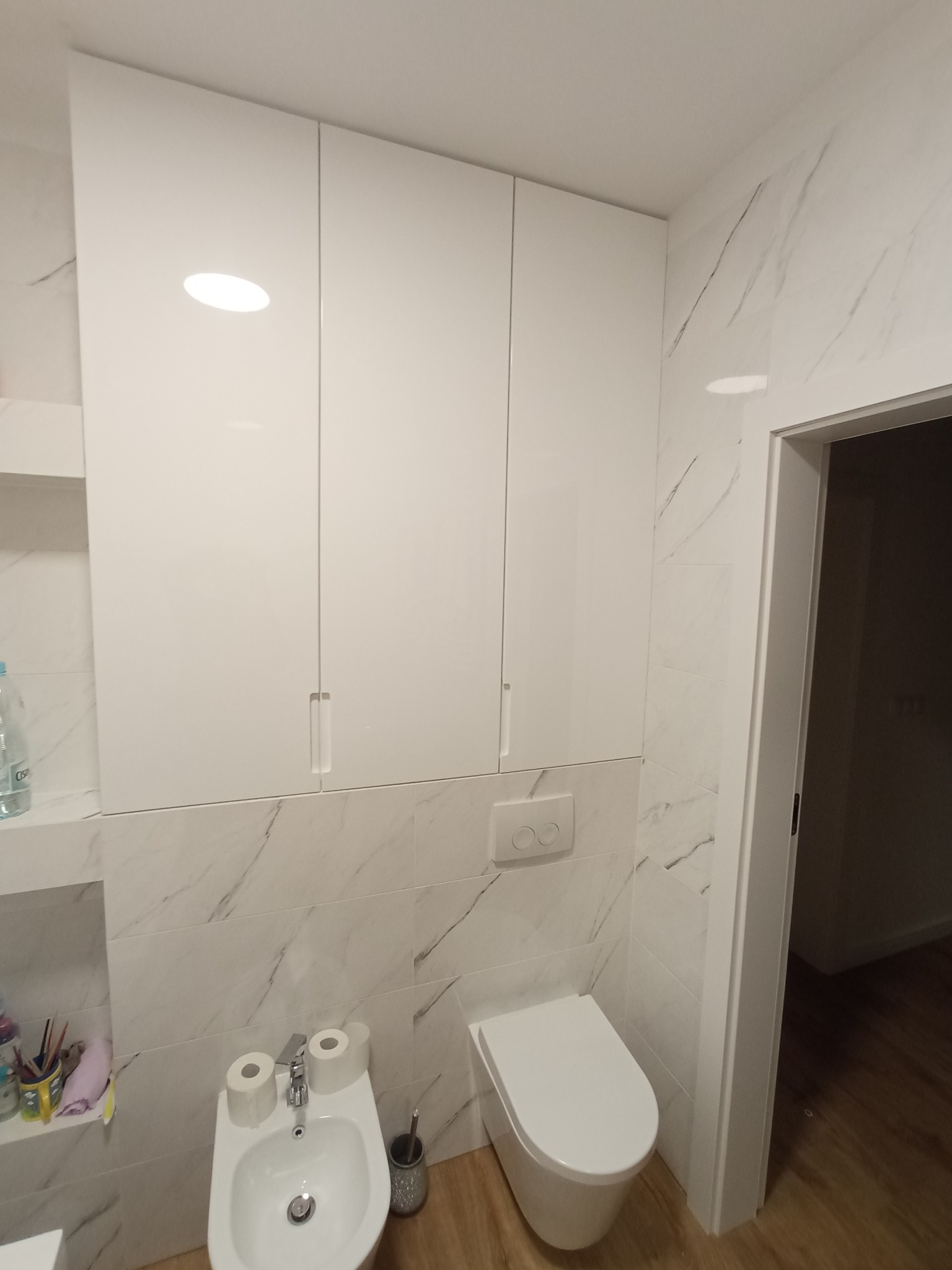 Nowoczesna łazienka z białą zabudową meblową pod sufit, marmurowe płytki na ścianach, wisząca toaleta i bidet, drewniana podłoga.