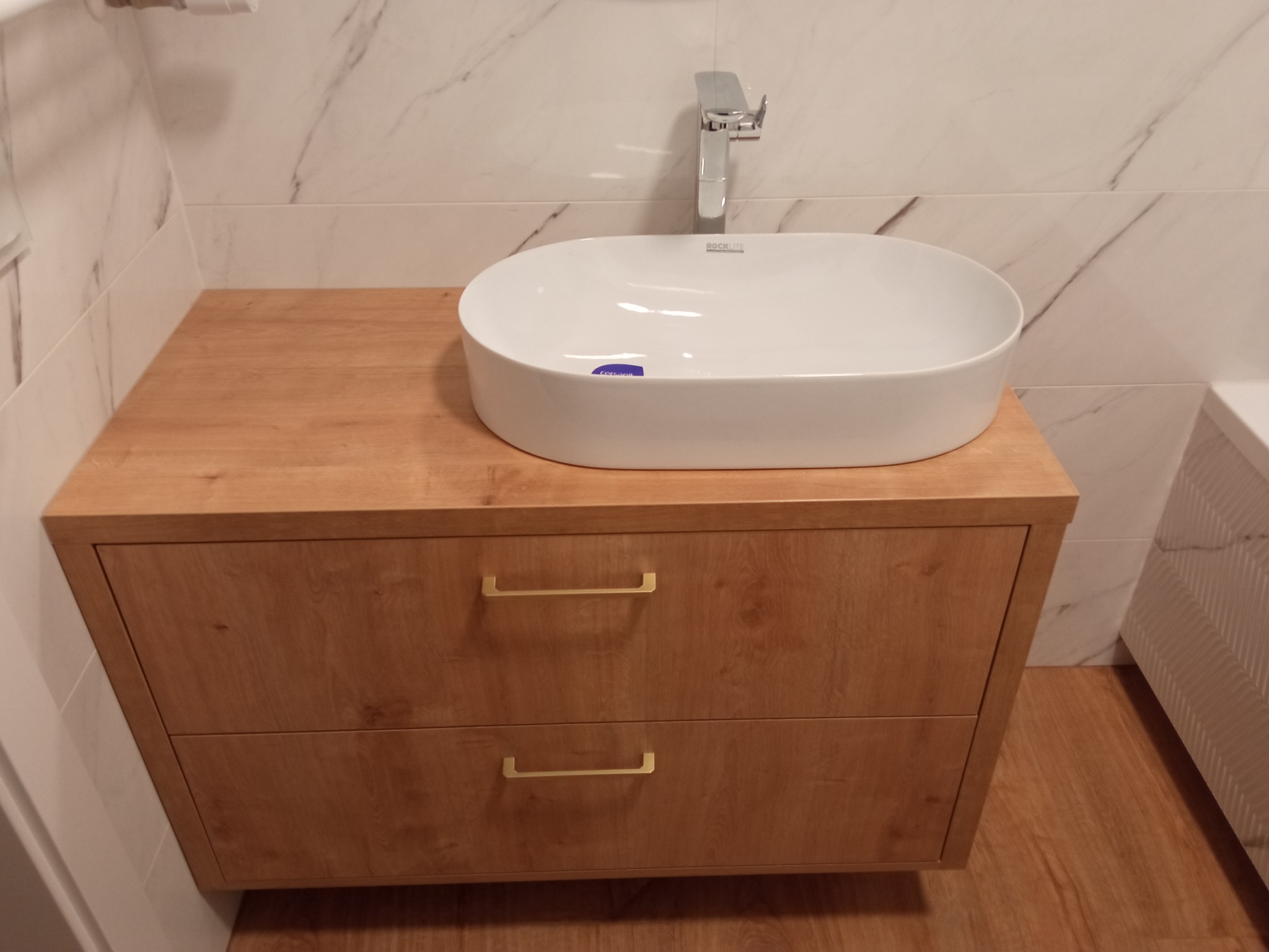 Łazienkowa szafka pod umywalkę z dwoma szufladami o drewnianym wykończeniu, biała umywalka nablatowa i chromowana bateria na tle marmurowej ściany.