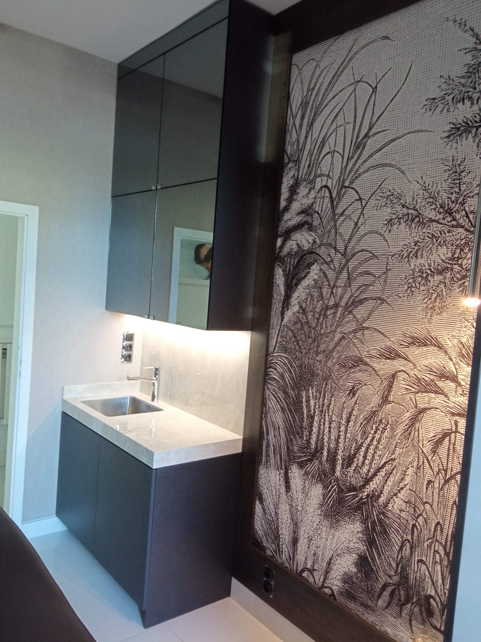 Nowoczesna łazienka z ciemnymi szafkami i blatem z jasnego marmuru, nad zlewem lustrzane szafki wiszące, na ścianie dekoracyjna tapeta w stylu retro z motywem roślinnym.