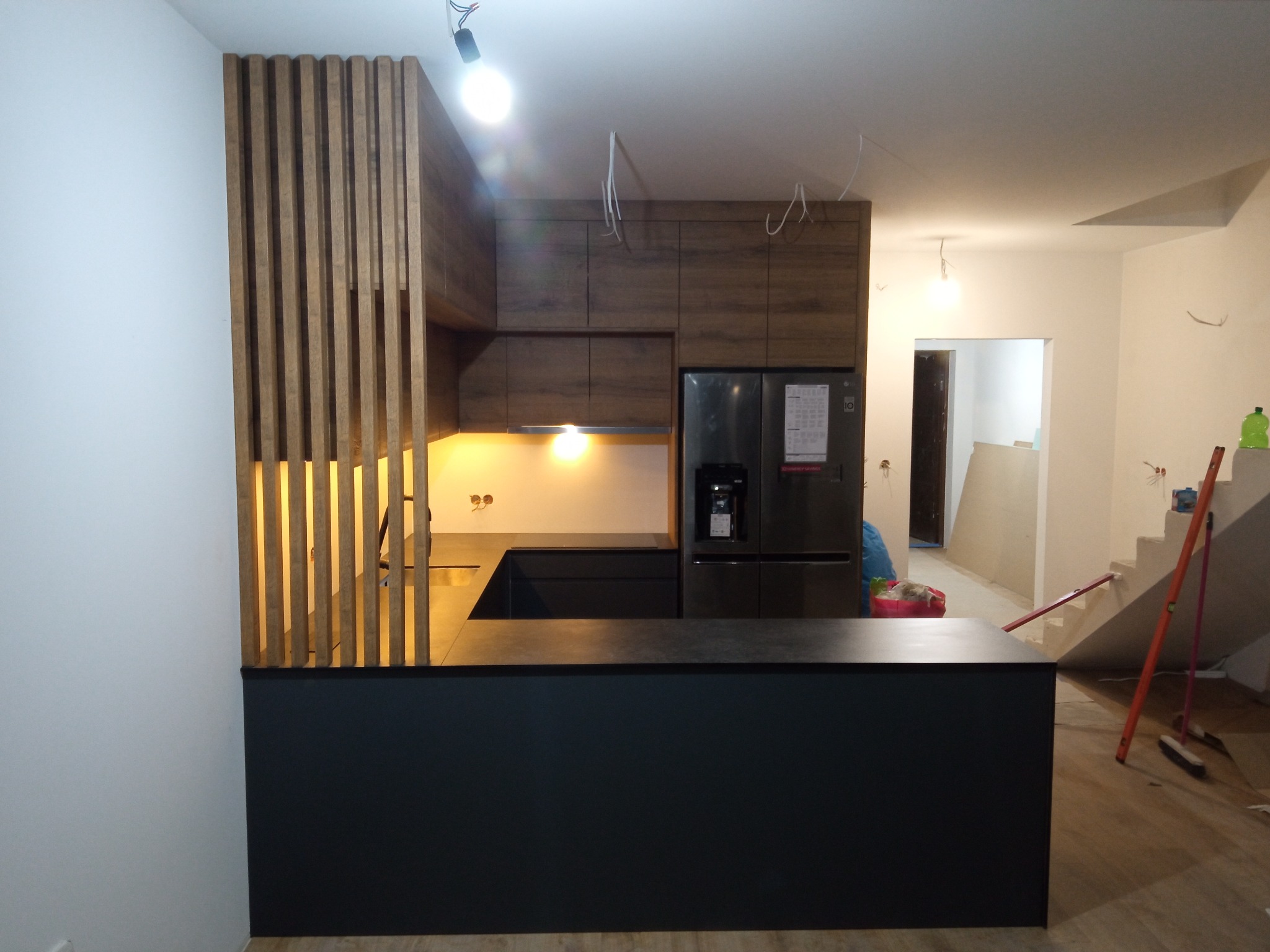 Częściowo ukończona, nowoczesna kuchnia z drewnianymi lamelami, czarnym blatem i zabudową w odcieniach brązu, widoczne kable wystające z sufitu i niedokończone schody w tle.
